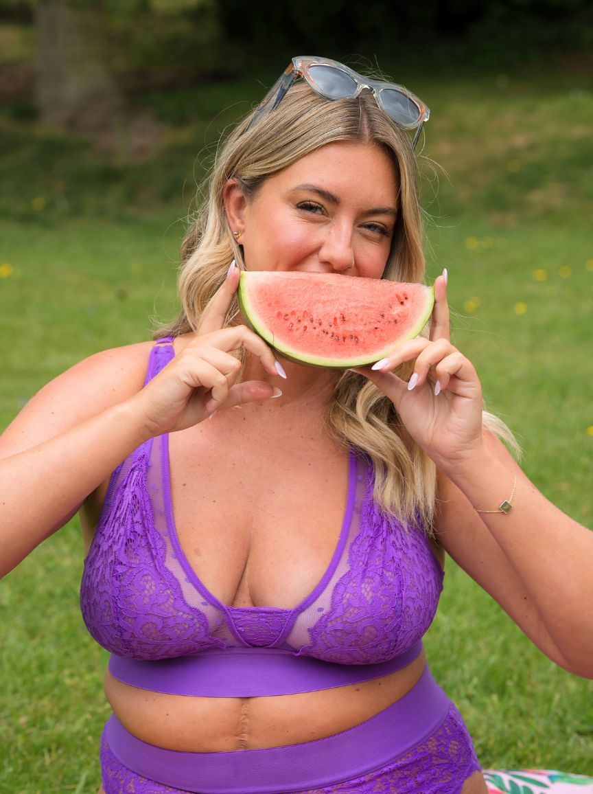 Blonde model smiling while wearing the Hallie Passion Purple lace bralette by Tutti Rouge, holding a slice of watermelon outdoors.